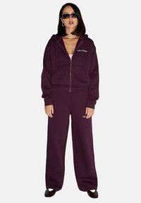Fioletowa wiatrówka na zamek z przednimi kieszeniami, połączona z dopasowanymi, luźnymi spodniami. Posiada teksturowany materiał i detal z logo na spodniach.