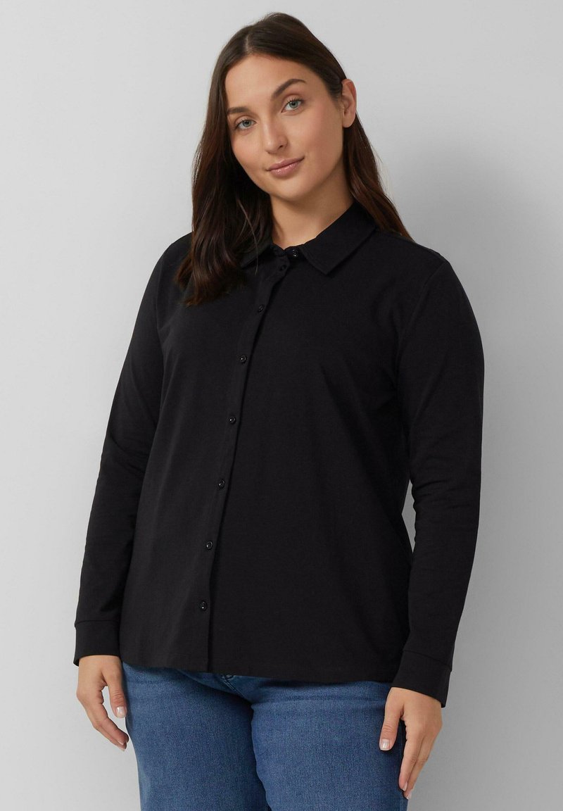Zwarte langemouw shirt gemaakt van zachte stof, met een puntige kraag en een knopen sluiting aan de voorkant, gecombineerd met blauwe spijkerbroek.