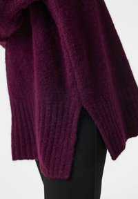 Gros plan d'un pull en maille violet foncé avec ourlet côtelé et fente sur le côté, porté sur un pantalon noir sur fond blanc.