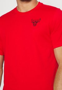 Rotes Baumwoll-T-Shirt mit einem schwarzen gestickten Bullenlogo auf der linken Brust. Kurze Ärmel und Crew-Neck-Design; glatte Textur.