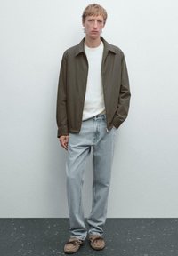 Oliven grøn zip-up jakke over hvid skjorte; lyseblå jeans; brune mokkasin-stil sko; glat stof med en afslappet pasform.