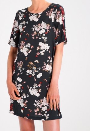 Femme portant une robe noire à manches courtes avec des motifs floraux blancs, roses et rouges et un col rond, debout devant un fond uni.