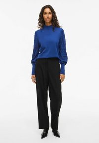 Blauer Strickpullover mit strukturierten Puffärmeln, kombiniert mit hochtaillierten schwarzen Hosen und spitzen schwarzen Absatzschuhen. Einfaches und elegantes Design.
