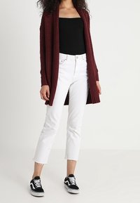 Burgundyröd stickad cardigan med öppen front, bärs över en svart topp, tillsammans med vita cropped jeans och svarta sneakers.