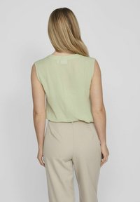 Blusa senza maniche di colore verde chiaro con una consistenza morbida, scollatura rotonda e una vestibilità leggermente ampia, abbinata a pantaloni beige su misura.