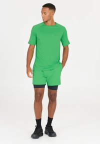Groene sportshirt met korte mouwen, gecombineerd met bijpassende groene shorts en zwarte compressiekorte broek eronder. Zwarte sportschoenen maken de look compleet.