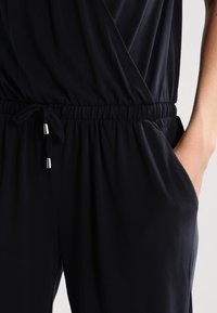 Marinblå jumpsuit med omslagdesign, elastisk midjeband och snörning. Innehåller sidfickor och glänsande silverdetaljer.