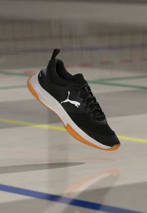 Chaussure de sport noire avec logo Puma blanc, semelle orange et semelle intermédiaire blanche, flottant au-dessus d'un sol de terrain de sport avec des lignes de terrain colorées.