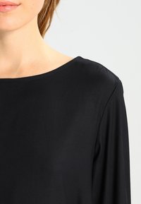 Haut noir avec un large col, des manches longues et un tissu lisse et léger avec un subtil brillant. Design simple et sans ornements.