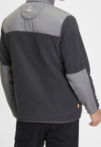 Grijze fleecejas met een gladde grijze bovenkant, hoge kraag en een oranje accentenlabel. Heeft een gestructureerde stof met een logo op de achterkant.