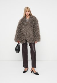 Χνουδιαστό καφέ παλτό με υφασμάτινη υφή, φορεμένο πάνω από γκρι μπλούζα; συνδυασμένο με σκούρες καφέ ψεύτικες δερμάτινες παντελόνες και μαύρες γόβες με λουράκι στη φτέρνα.