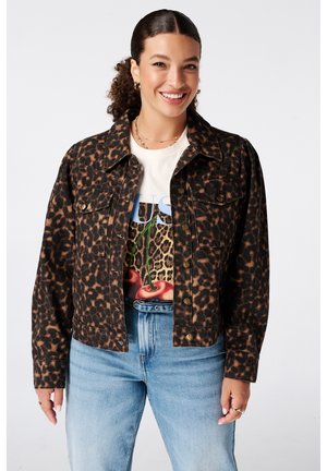 Femme souriante aux cheveux bouclés porte une veste à imprimé léopard sur un t-shirt graphique cerise et léopard, associée à un jean bleu clair.