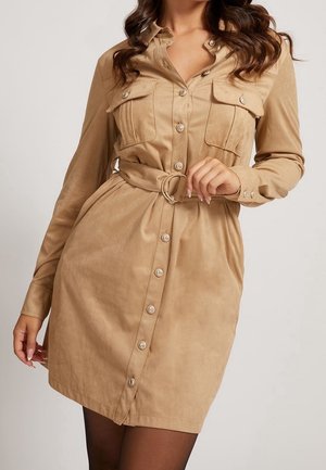 Mujer que lleva un vestido camisa marrón claro con botones, mangas largas, bolsillos en el pecho y un cinturón a juego con una hebilla redonda.