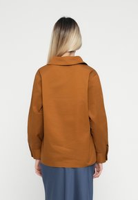 Person med langt blondt hår iført en brun langærmet skjorte og nederdel i blå, vendt væk mod en ensartet hvid baggrund.