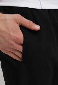 Černé kalhoty s hladkým povrchem, které mají boční kapsu. Ruka spočívá v kapse, což ukazuje na střih a designové detaily kalhot.