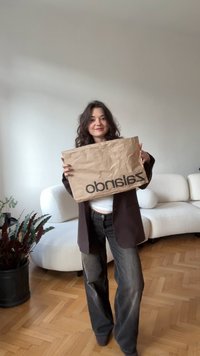 Busta di carta marrone con testo nero che recita "zara", tenuta da una persona in blazer scuro e jeans. Sfondo chiaro e ambiente interno.