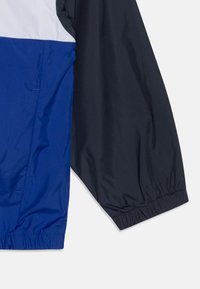 Rompevientos azul y marino con un acento blanco, que presenta una textura ligera y suave, además de puños elásticos. Incluye dos bolsillos laterales.
