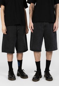 Shorts en denim noir avec une coupe ample, des poches latérales et un ourlet brut. Associés à des chaussettes noires et des chaussures lacées noires brillantes.