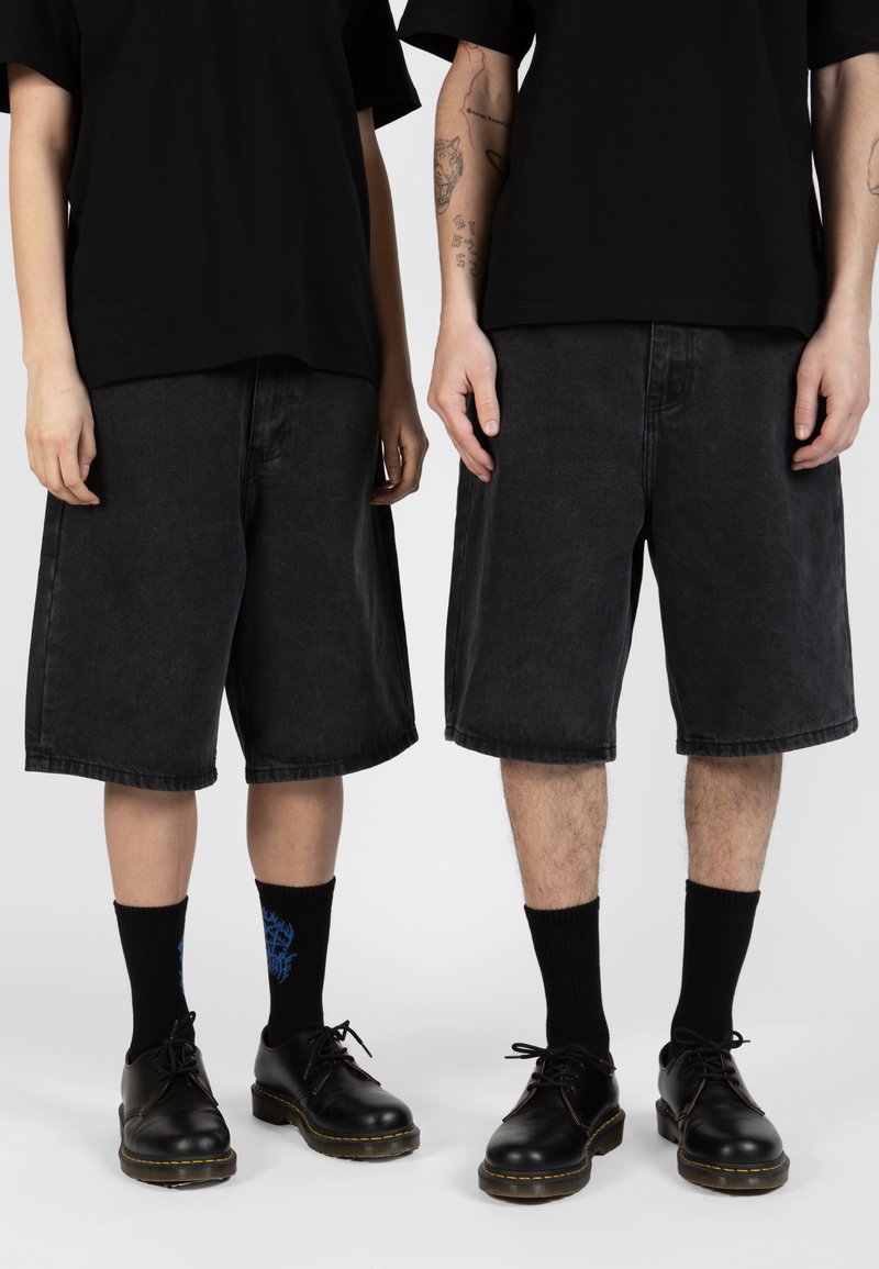 Shorts en denim noir avec une coupe ample, des poches latérales et un ourlet brut. Associés à des chaussettes noires et des chaussures lacées noires brillantes.