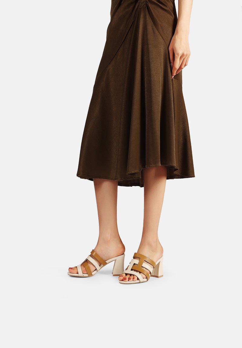Sandali con tacco a blocco, con cinturini marroni e beige, caratterizzati da un design geometrico e con il retro aperto, abbinati a una gonna fluida marrone lunga fino alla caviglia.