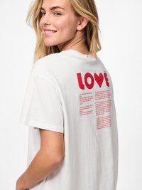 Nasmejana žena sa plavom kosom u bijeloj majici s velikim crvenim natpisom "LOVE" s srcem i pjesmom na leđima.