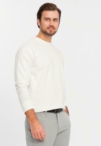 Mann mit Bart, der ein weißes Langarmhemd und hellgraue Hosen trägt, steht mit einer Hand in der Tasche vor einem weißen Hintergrund.