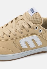 Beige sneakers with a suede finish, round toe, tan laces, mesh tongue, and a white logo design. Rubber sole with a textured grip.
