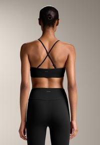 Črn športni modrček s kravato čez hrbet, v kombinaciji s črnimi hlačami z visokim pasom. Obe oblačili imata gladko teksturo in diskretno blagovno znako.