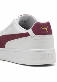 Adidaș alb cu o dâră maron, partea superioară din piele texturată, guler căptușit, talpă din cauciuc și logo auriu pe călcâi.