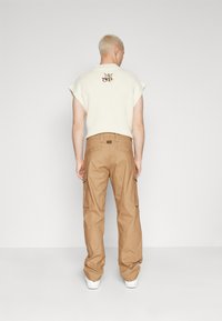 Lys beige strikket topp med korte ermer; bakdelen har et brodert logo. Brune cargobukser med sidelommer; hvite sneakers.