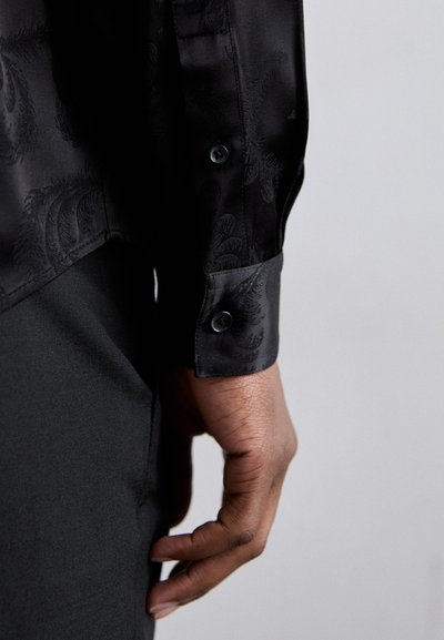 Camicia nera in satin a maniche lunghe con motivo paisley, dotata di polsini abbottonati e texture liscia. Mano appoggiata su un tessuto nero.