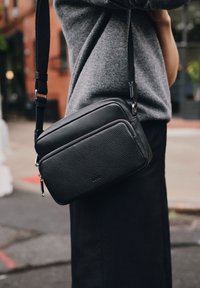 Svart läder crossbody-väska med texturerad yta, rektangulär form, blixtlås och justerbar rem. Konturerad söm-detalj.