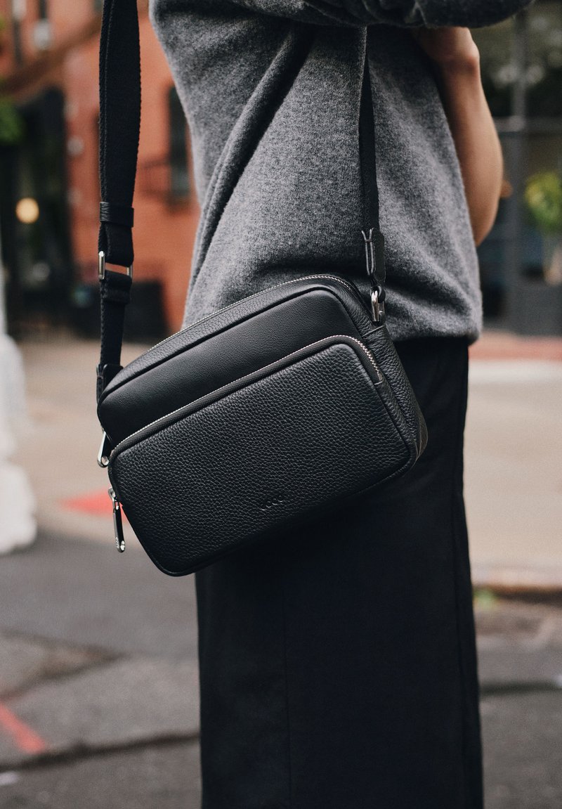 Svart läder crossbody-väska med texturerad yta, rektangulär form, blixtlås och justerbar rem. Konturerad söm-detalj.