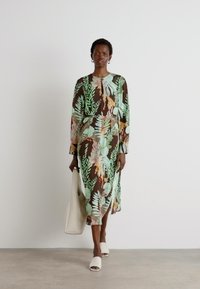 Abito lungo a maniche lunghe, marrone, con motivi floreali verde e arancione, stoffa fluida, spacco laterale, abbinato a scarpe bianche con punta aperta e una borsa beige.