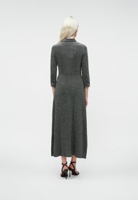 Moteris su ilgu šviesiais plaukais, vilkinti pilką ilgu rankovėmis midi suknelę ir juodas aukštakulnes sandalus, stovi nugara į kamerą ant baltos fono.