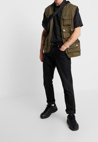 Grüne multifunktionale Weste mit mehreren Taschen über einem schwarz-karrierten Hemd. Schwarze Jeans und klobige schwarze Sneaker runden das Outfit ab.