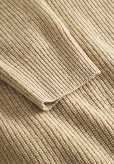 Bežani rebrasti pleteni materijal s mekim teksturom, s preklopljenim rubom. Uzorak se sastoji od okomitih linija, stvarajući slojeviti izgled.