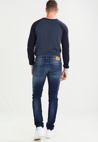 Mörkblå jeans med slim fit, ljusblå sömmar och bakfickor; matchat med en marinblå sweatshirt och vita sneakers.