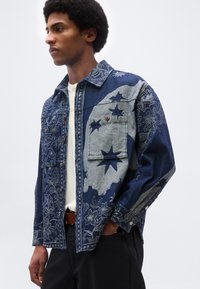 Άνδρας με μπλε τζιν μπουφάν με αστέρια και λουλουδάτα σχέδια, λευκό πουκάμισο από μέσα, μαύρο παντελόνι και καφέ ζώνη, με τα χέρια στις τσέπες.