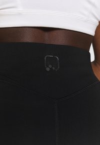 Zwarte high-waisted leggings met een gladde textuur. Heeft een subtiele logo-detail nabij de tailleband en zichtbare stiksels langs de naden.
