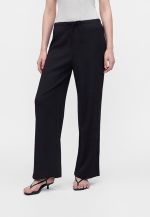 Mujer que lleva pantalones negros de pierna ancha con cinturilla ajustable y sandalias negras de tacón sobre un fondo blanco.