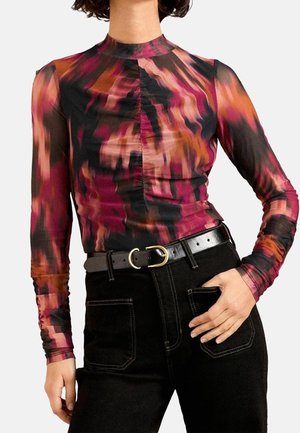 Frau trägt ein eng anliegendes Langarmshirt mit hohem Kragen und einem lebhaften abstrakten Muster in Rot, Orange und Schwarz, kombiniert mit einer schwarzen Hose und einem Gürtel.