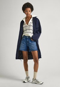 Veste légère marine avec cordon de serrage, haut en tricot crème avec tissage ouvert, shorts en denim, chaussettes beiges et chaussures de sport grises.