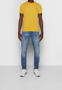 Žluté bavlněné tričko s červeným logem "TOMMY JEANS" na prsou, spárované s světle modrými džínami a bílými teniskami.