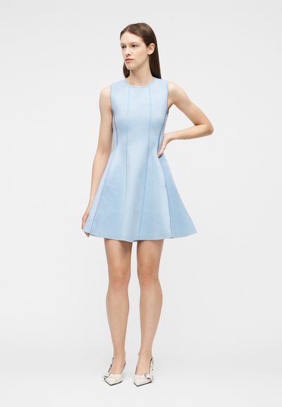 Femme debout, main gauche sur la hanche, portant une robe sans manches bleu clair en ligne A et des talons blancs à bout pointu, sur un fond uni.