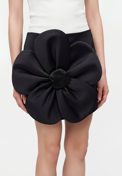 Mini-jupe noire avec une grande décoration rembourrée en forme de fleur couvrant le devant. Le mannequin porte un haut blanc sans manches.