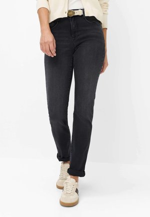 Person iført opsmøgede sorte jeans, hvide sneakers, en hvid top og en cremefarvet cardigan, stående mod en ensfarvet baggrund.