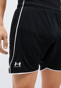 Κοντινό πλάνο ατόμου που φοράει μαύρο αθλητικό σορτς Under Armour με λευκή περίγραμμα και λογότυπο στο αριστερό μηρό.