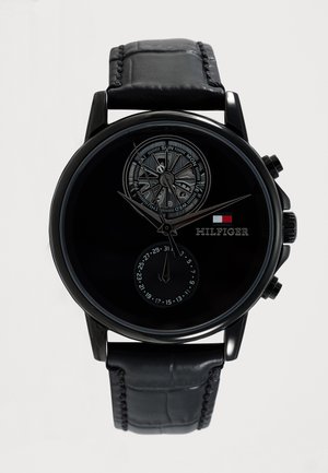 Ρολόι Tommy Hilfiger με μαύρο δερμάτινο λουράκι, μαύρο καντράν, υποκαντράν ημέρας και ημερομηνίας, ασημένιους δείκτες και μικρό κόκκινο, λευκό και μπλε λογότυπο.
