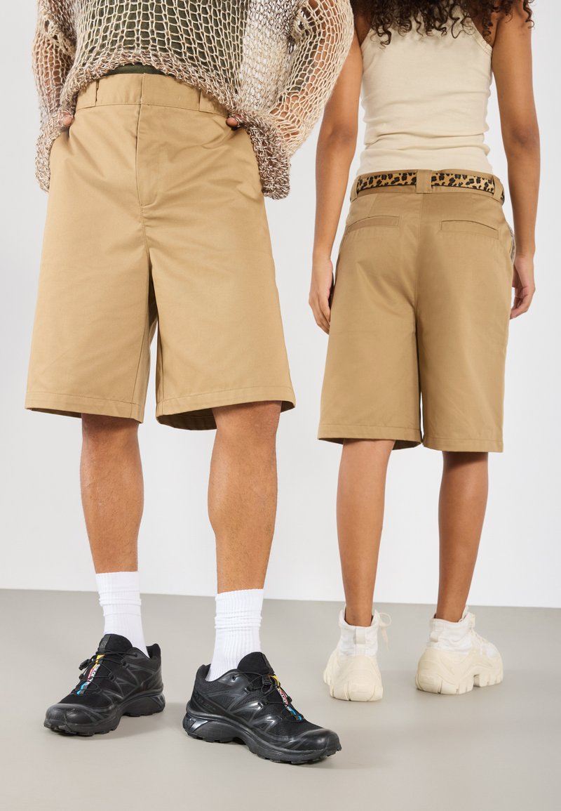 Shorts en coton beige avec des plis, présentant une ceinture avec un motif léopard marron sur une paire. Deux silhouettes sont présentées de dos, portant des chaussures différentes.